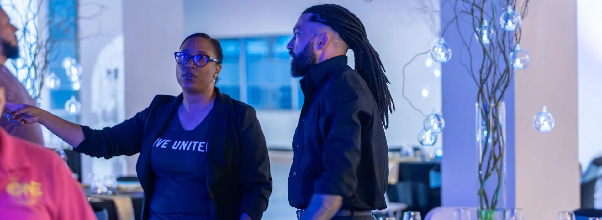 Woman in a black jacket and a bearded man in black talking beside a table with glass orb lighting and blue-tinted decor