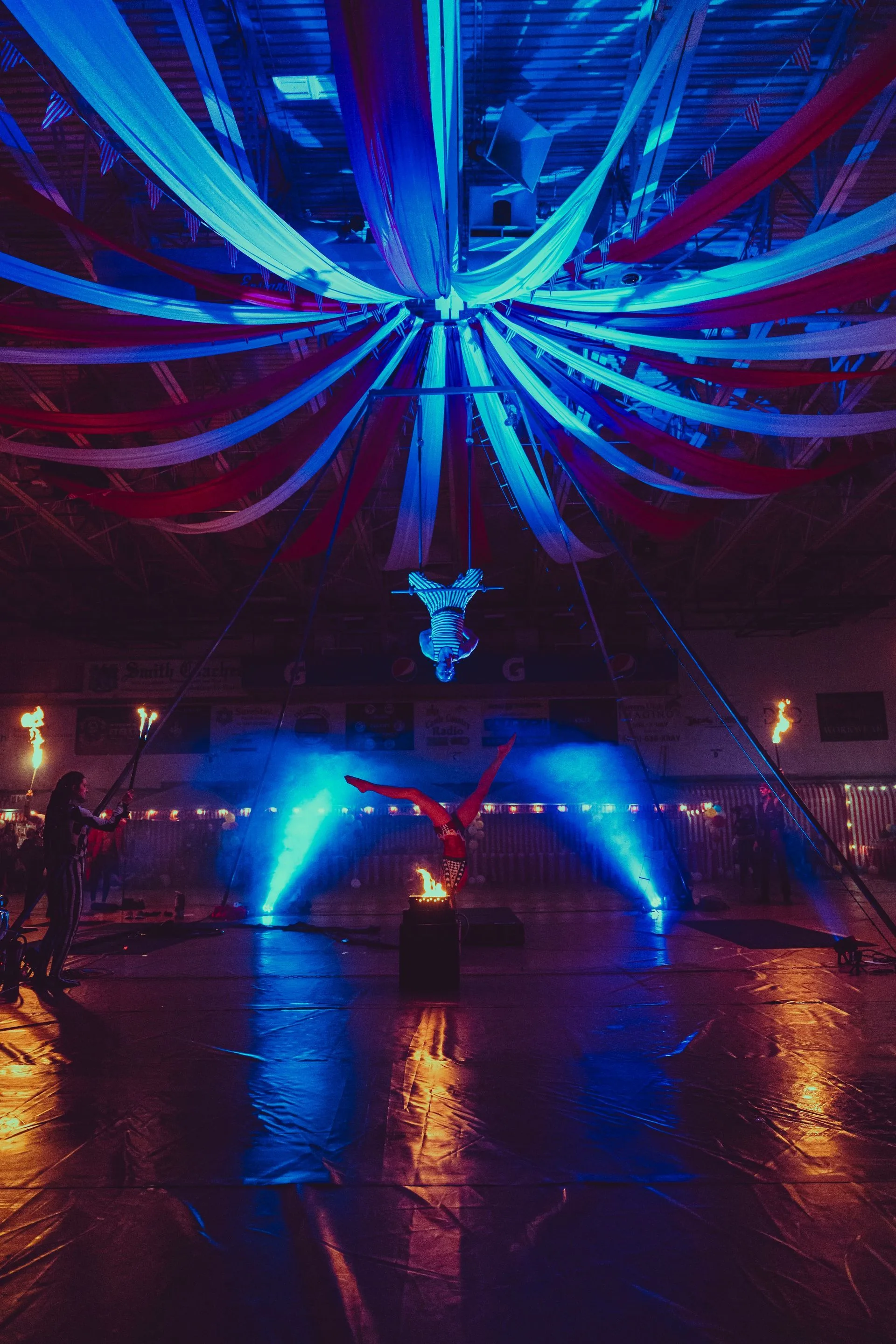 A person is performing aerial acrobatics in a dark room with blue and red ribbons hanging from the ceiling.