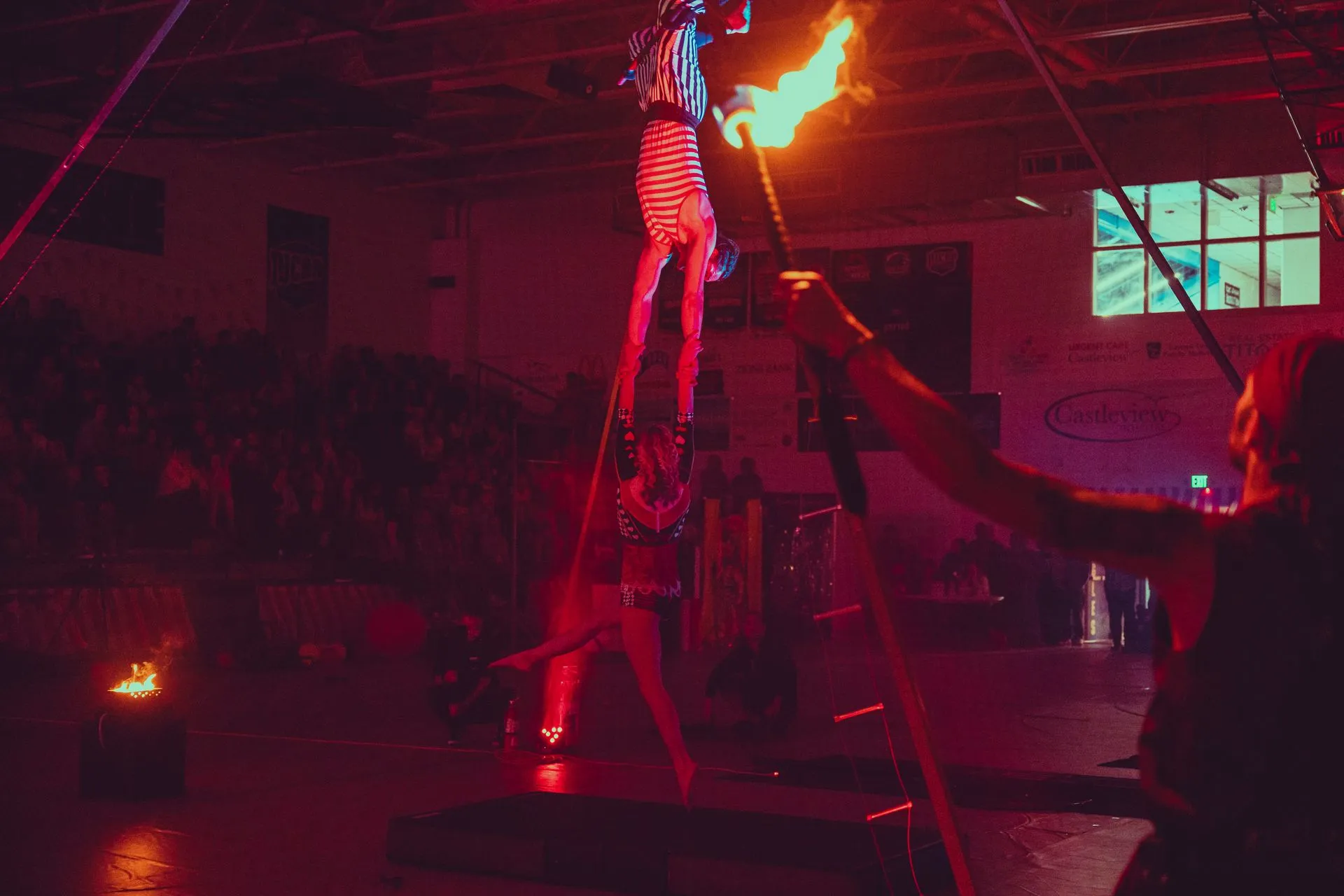 A man is holding a torch while a woman performs aerial acrobatics in a dark room.