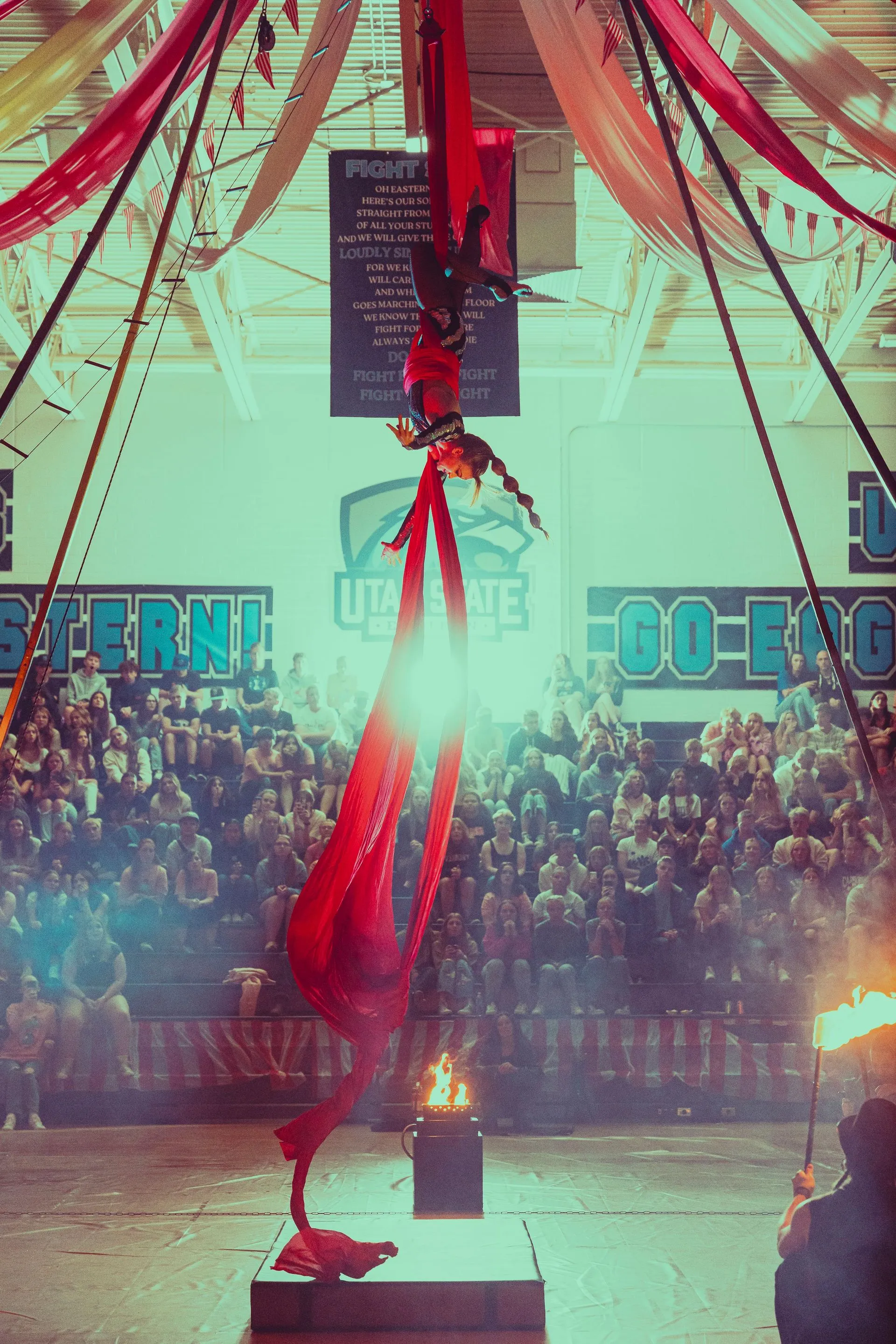 A person is hanging from a red aerial silk in front of a crowd.