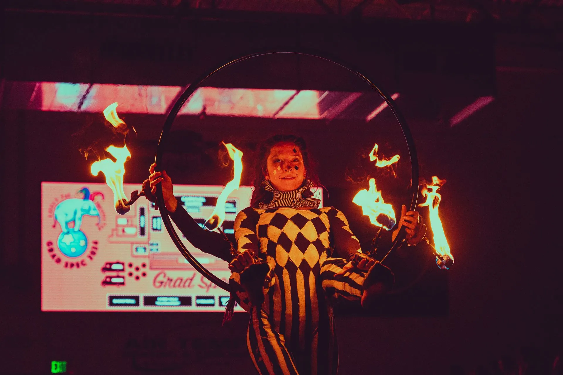 A man in a clown costume is holding a hula hoop with fire coming out of it.