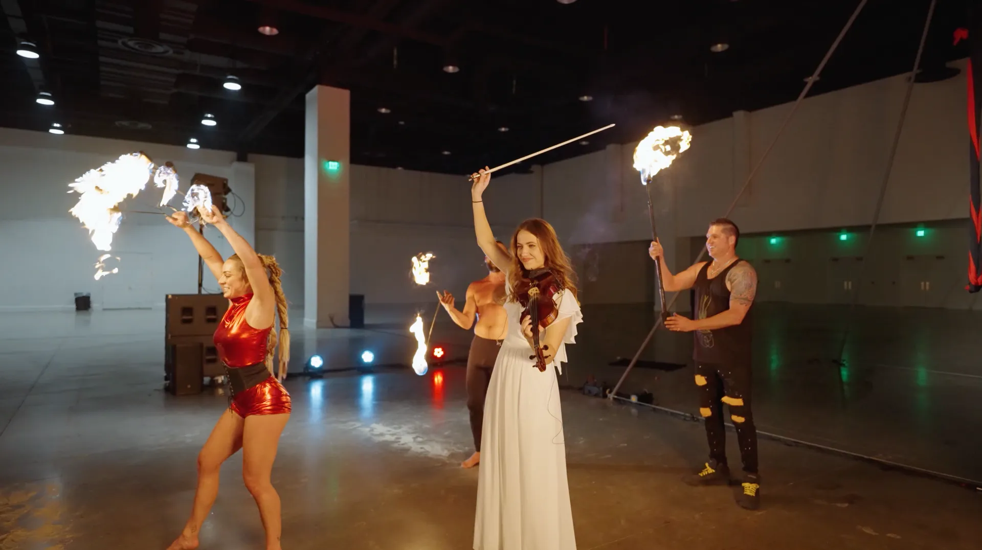 A group of people are performing a fire show in an empty room with a violinist.