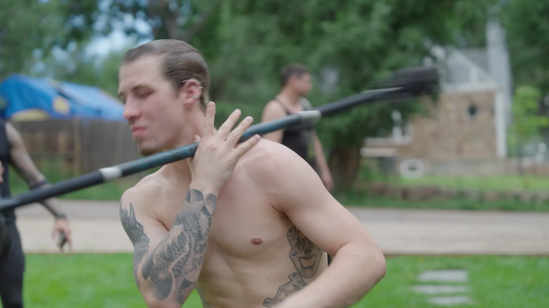 A shirtless man with a tattoo on his arm is holding a stick over his shoulder.