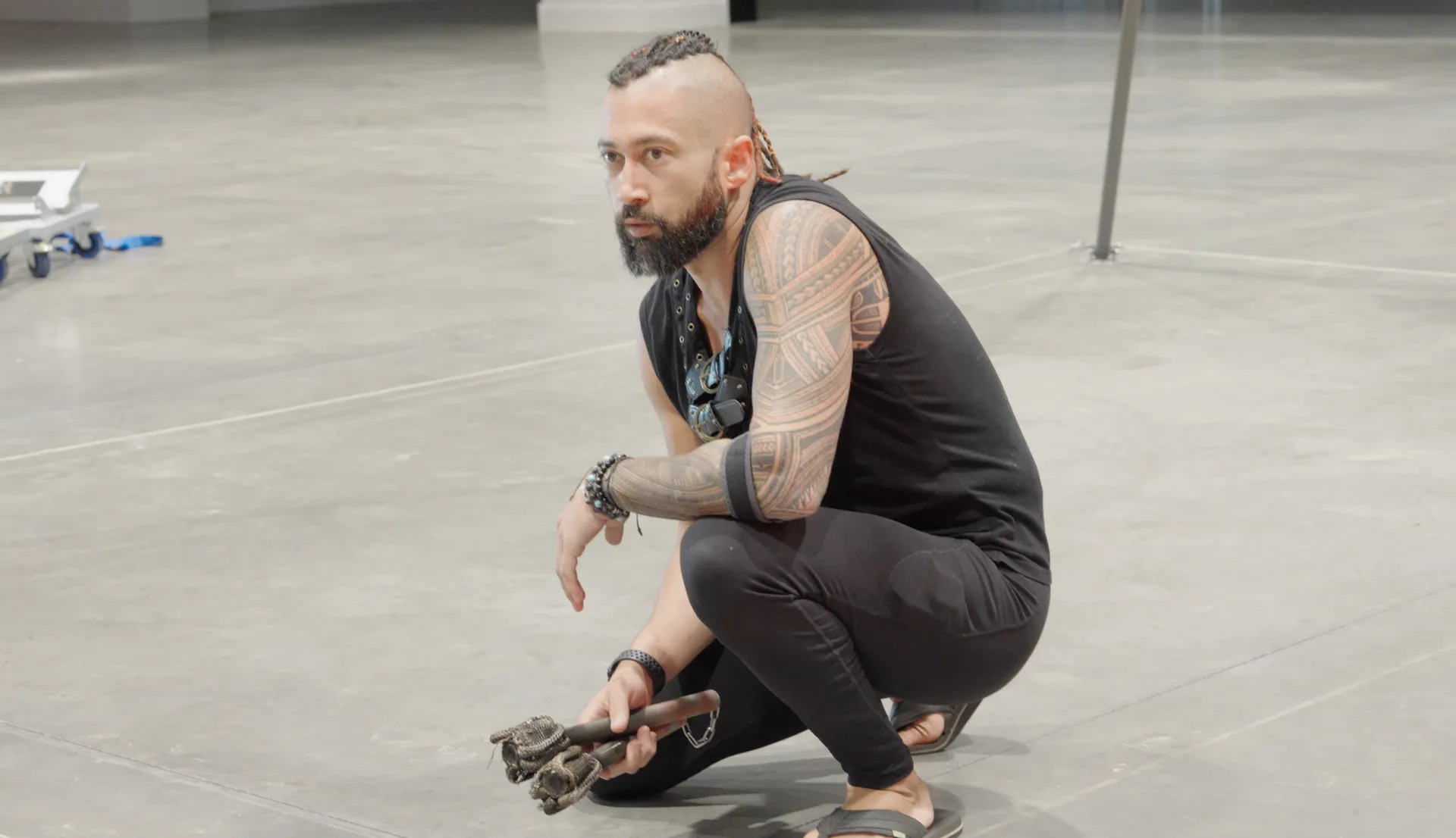 A man with a beard is squatting down on the floor holding a pair of roller skates.