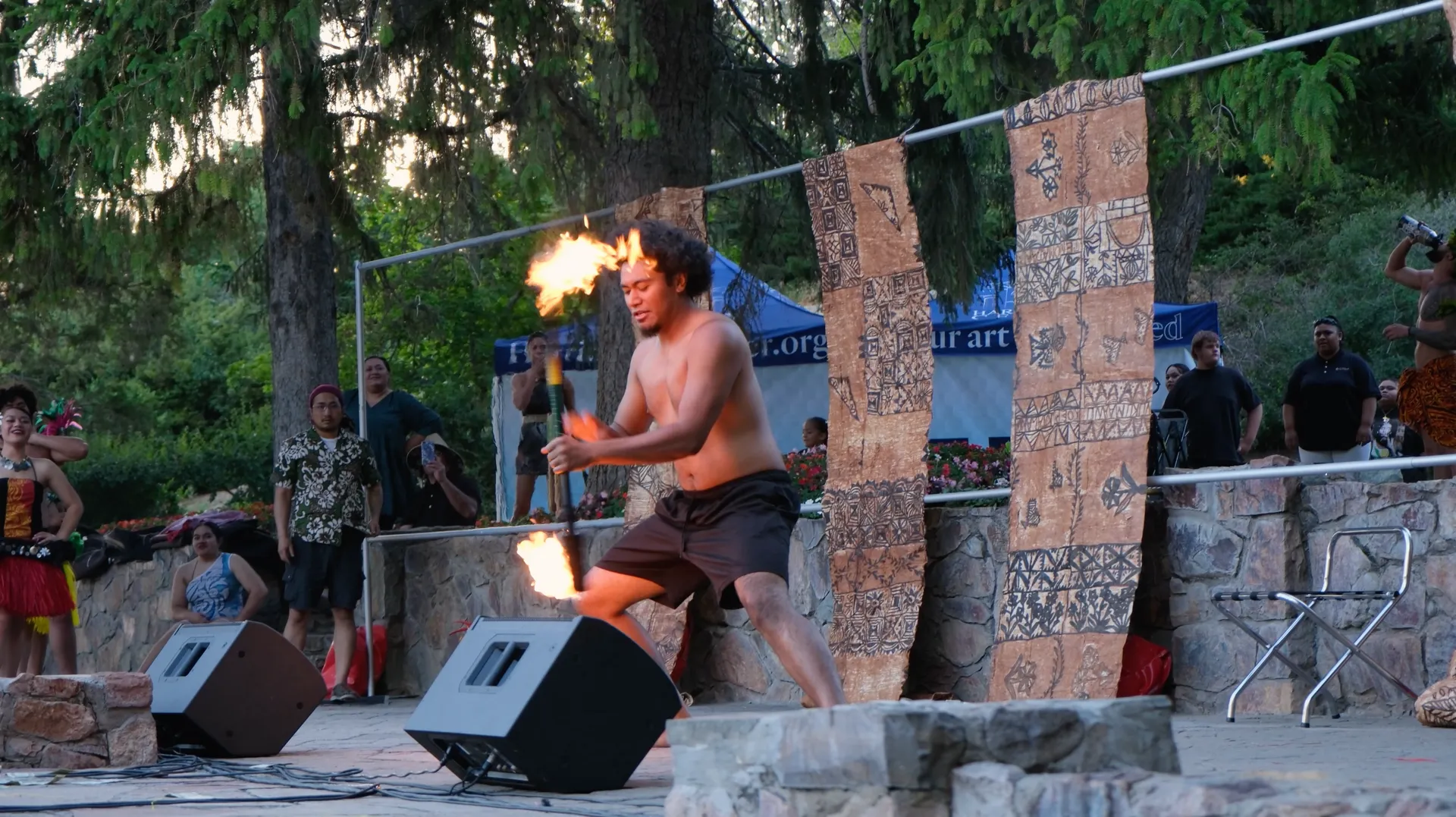A man without a shirt is holding a fire torch in front of two speakers.
