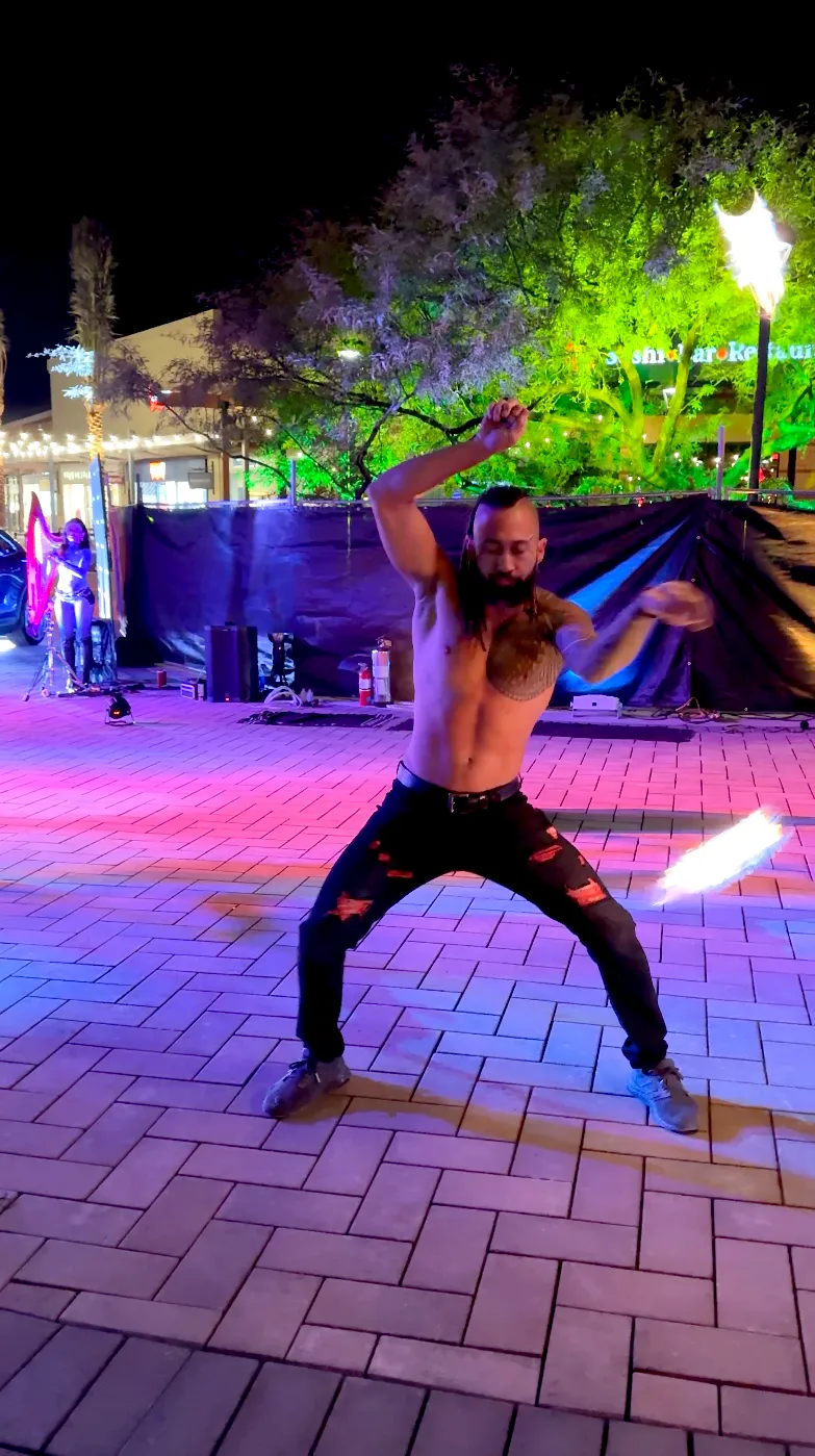 A shirtless man is playing a violin on a brick sidewalk at night.