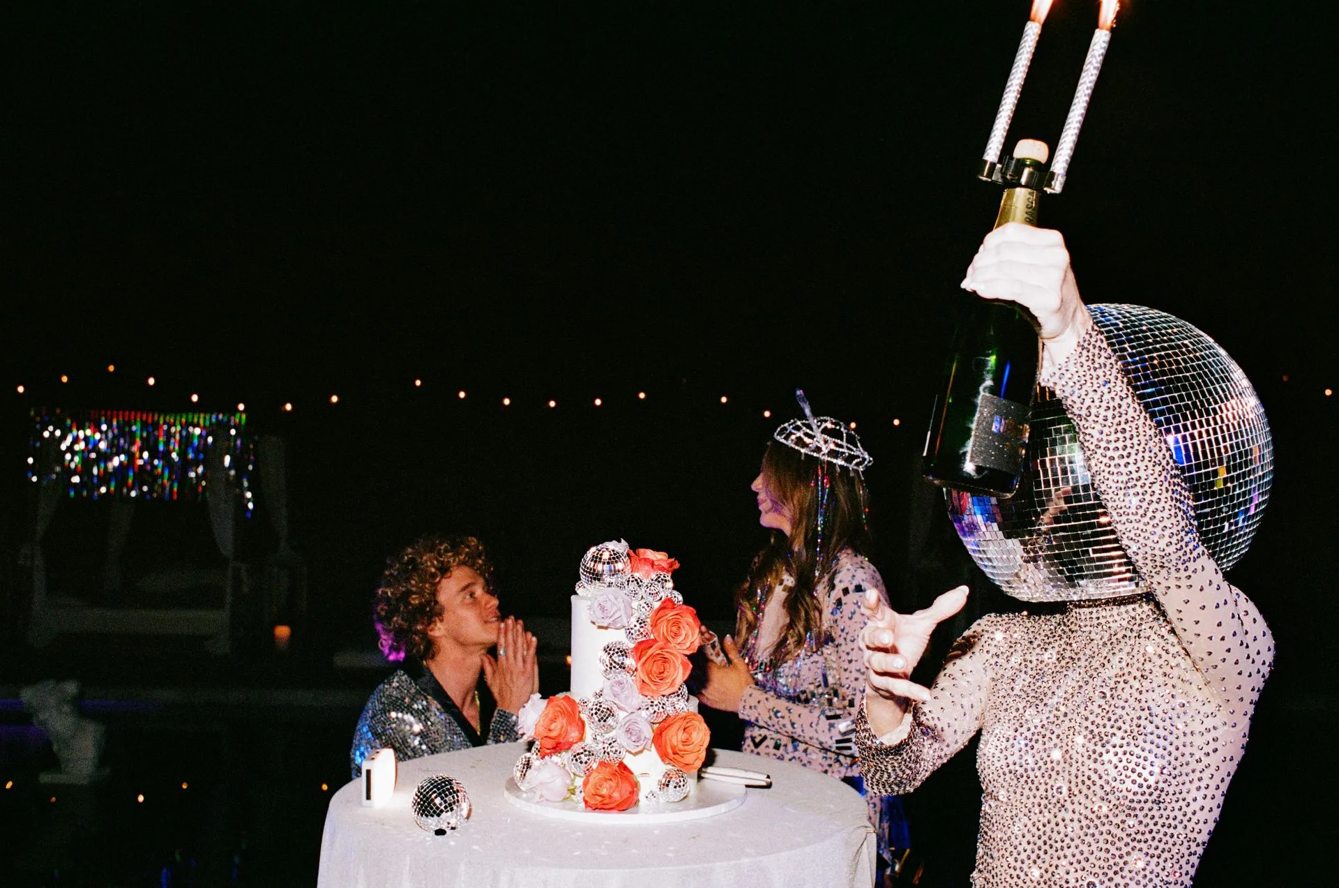 People celebrating outdoors at night, cake with orange flowers, disco ball suit, one holding a champagne bottle.
