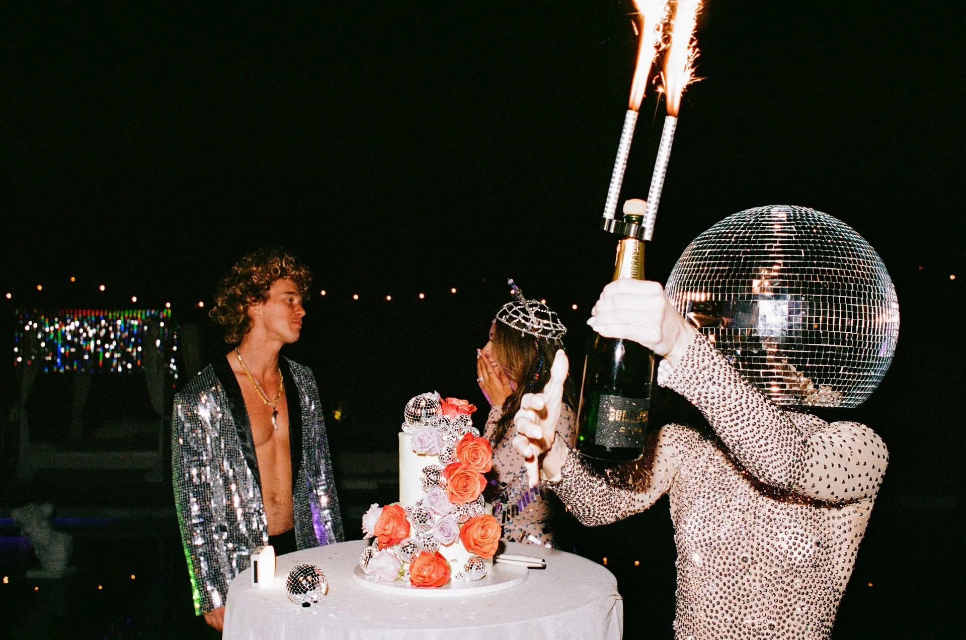 Person in disco ball head opens champagne with sparklers above a cake; man in sequin jacket watches.
