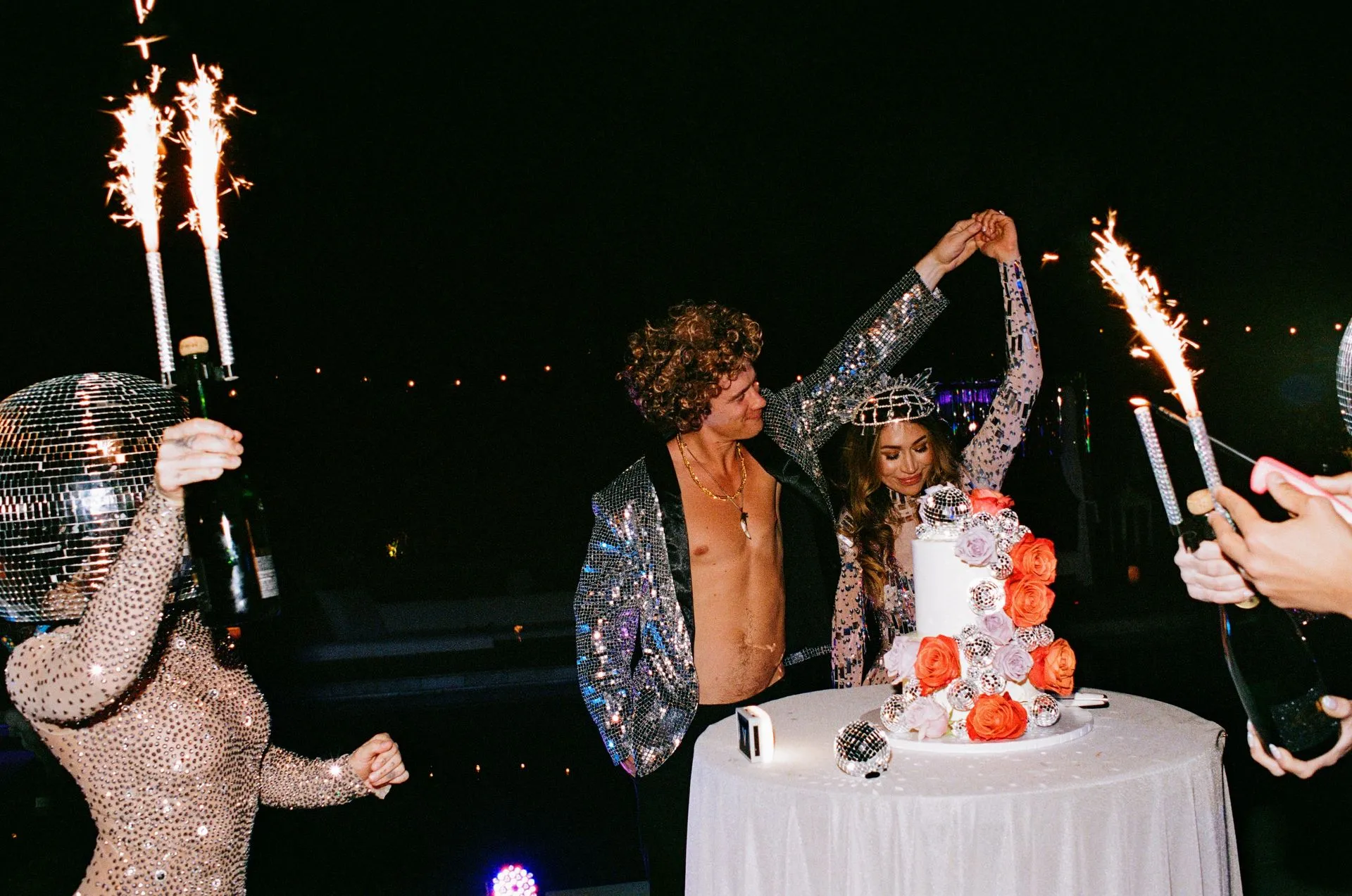 Couple celebrates next to a cake with sparklers. A person in a sequined outfit pours champagne. Nighttime setting.