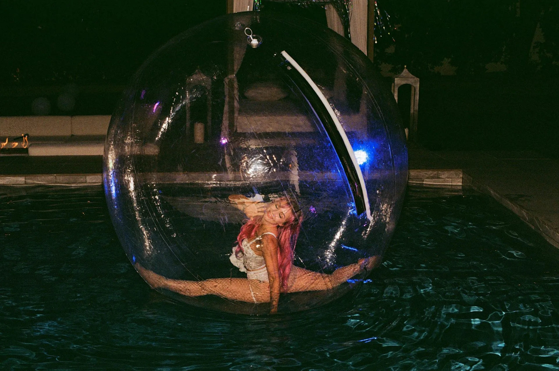 A person splits inside a clear orb in a pool, lit by blue and white lights.