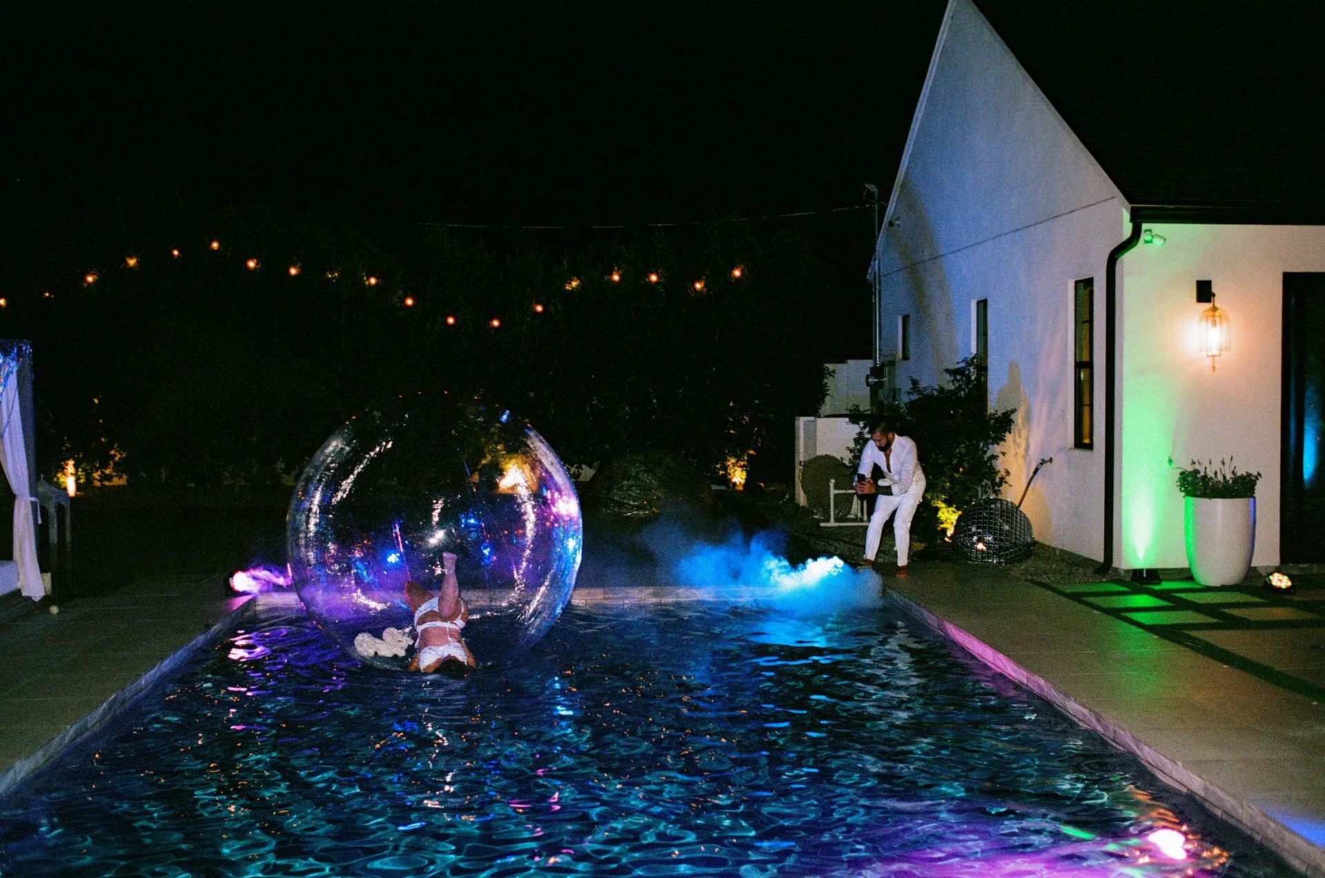 Person in a large, clear inflatable bubble in a pool at night. Colorful lights illuminate the water.