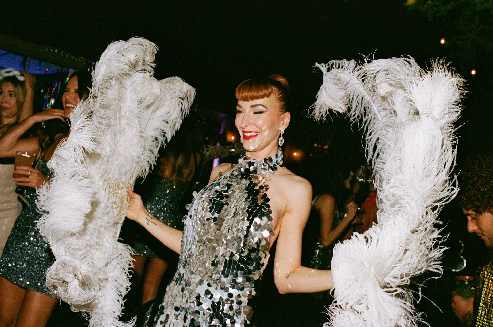 Woman in silver sequin dress dances at a party, holding feather boas. Others in background.