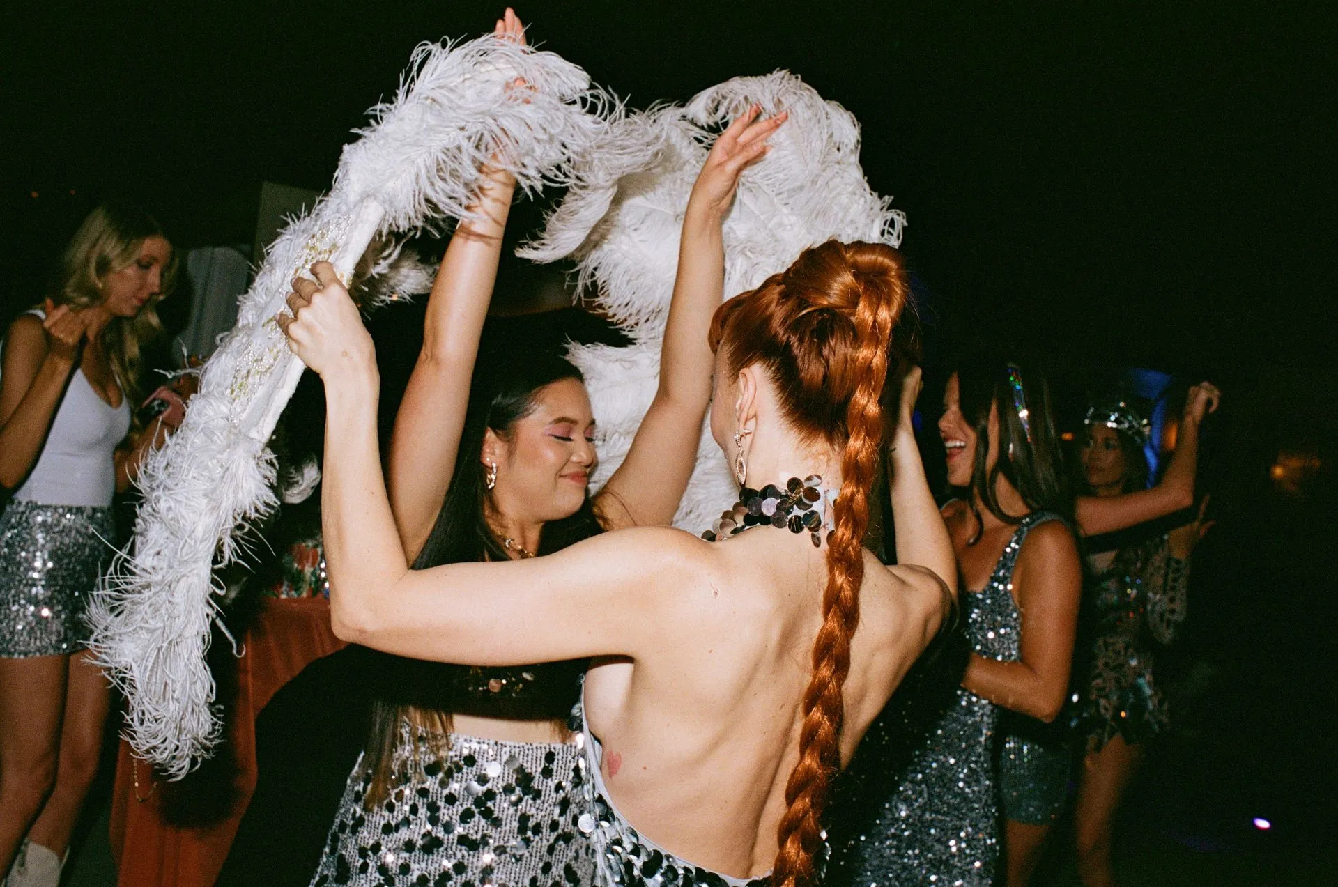 People dancing, one with long red braid holding feather boa, wearing silver sequin clothing at a party.