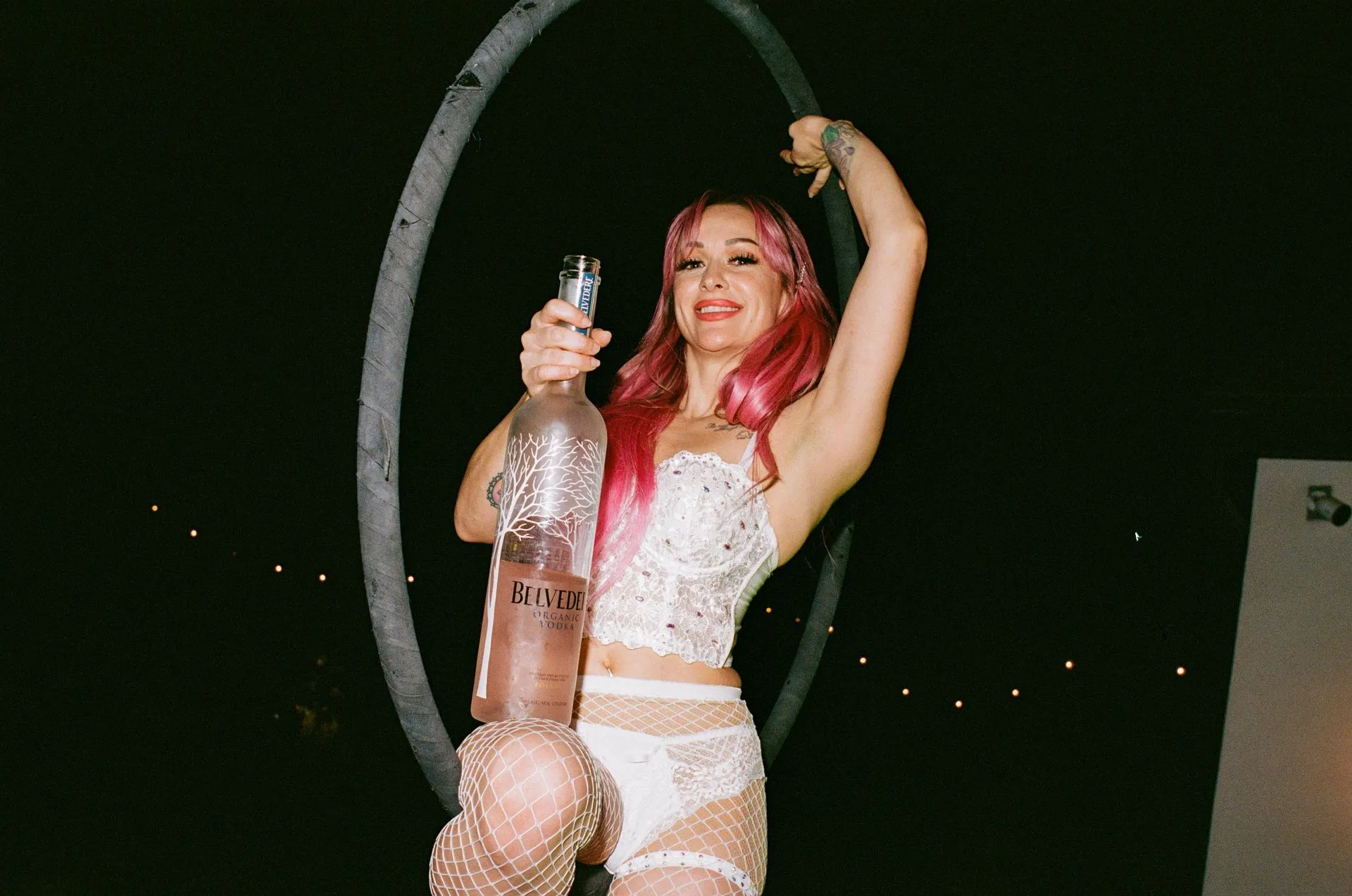 Woman in white outfit holding large vodka bottle, smiling, under a ring.