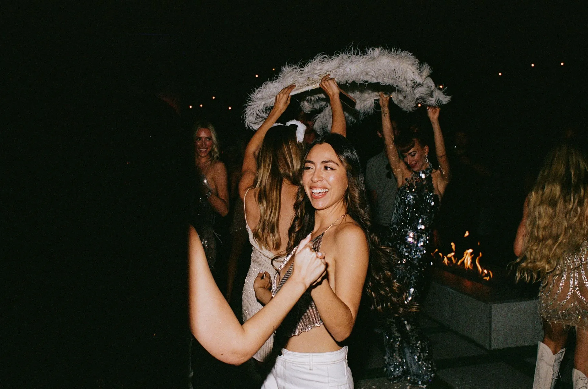 People dancing outdoors at night, smiling, with a feathered boa; bonfire in the background.