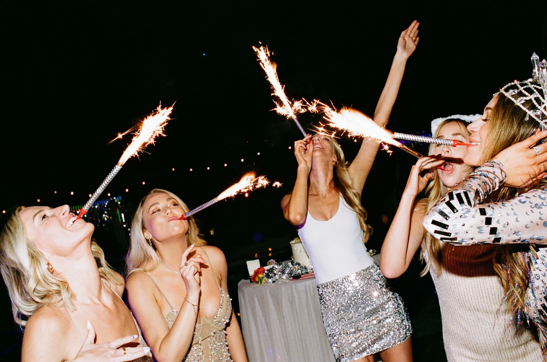 Five women at night, holding sparklers to their mouths. Celebrating, with raised arms and sparkly clothing.