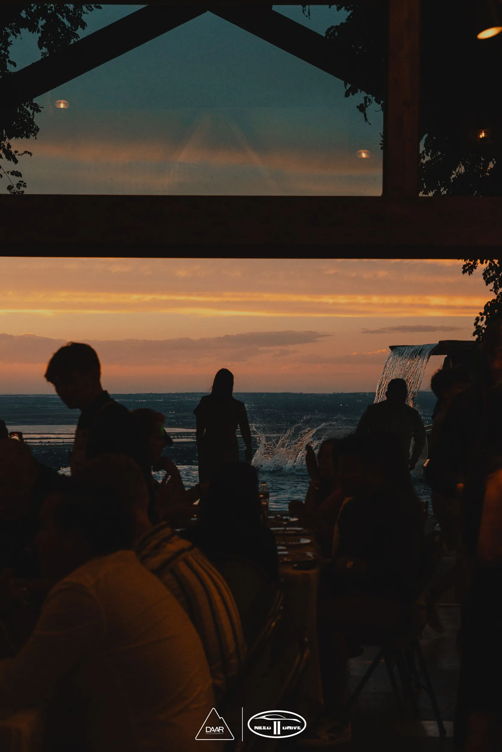 A group of people are standing in front of a window overlooking the ocean at sunset.