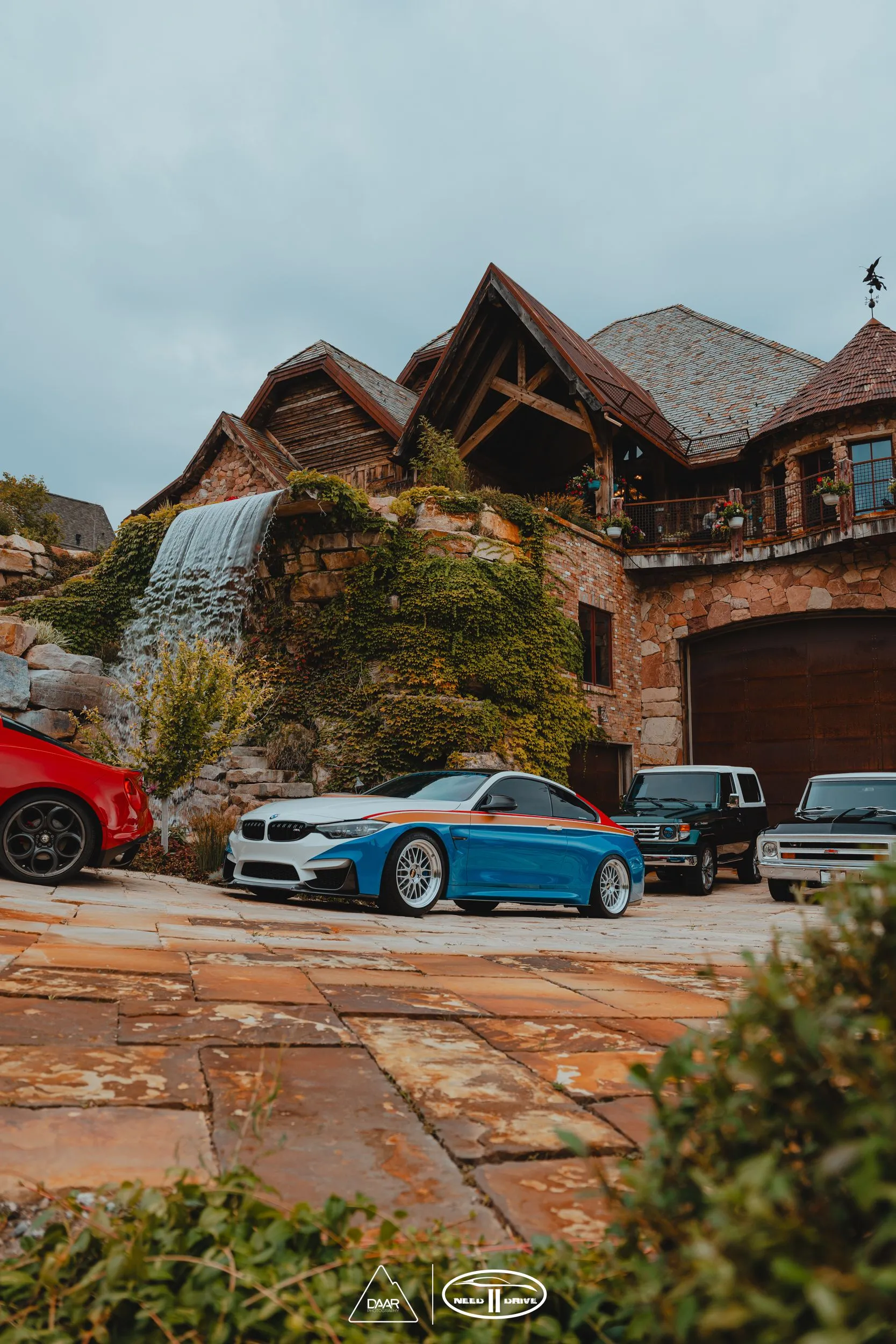 Three cars are parked in front of a large house with a waterfall in the background.