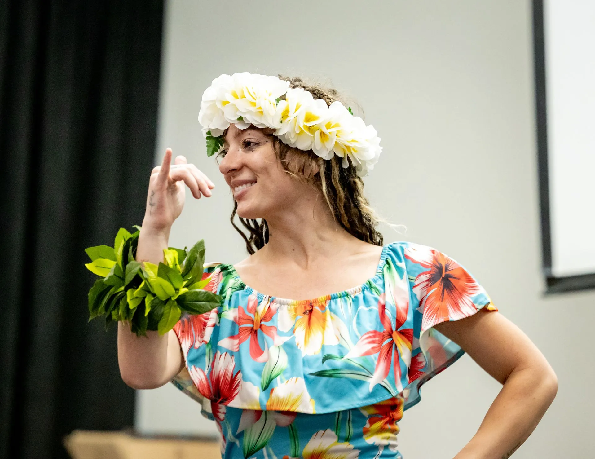 A woman wearing a floral dress and a flower crown is dancing a hula.
