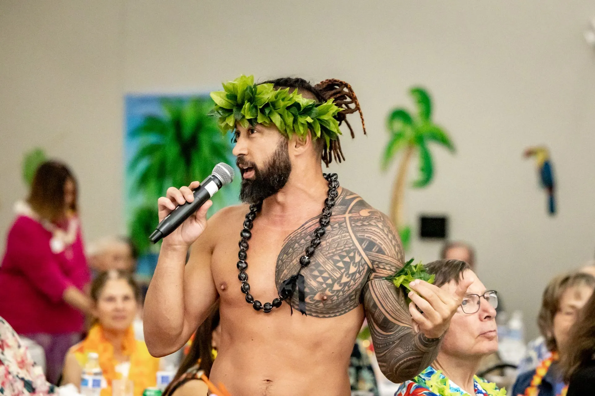 A shirtless man is singing into a microphone while wearing a lei.