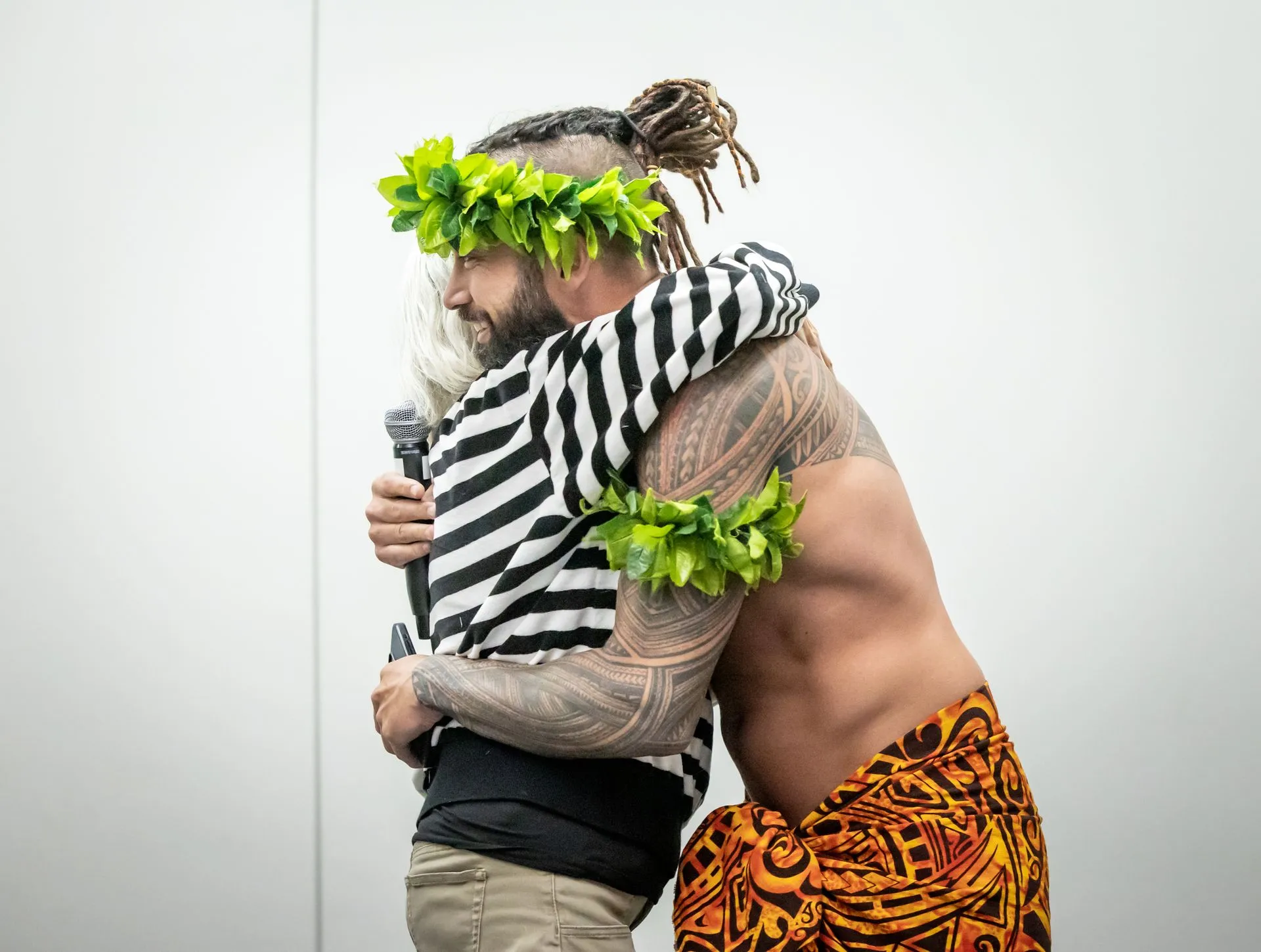 A man in a striped shirt is hugging another man in a hawaiian outfit.