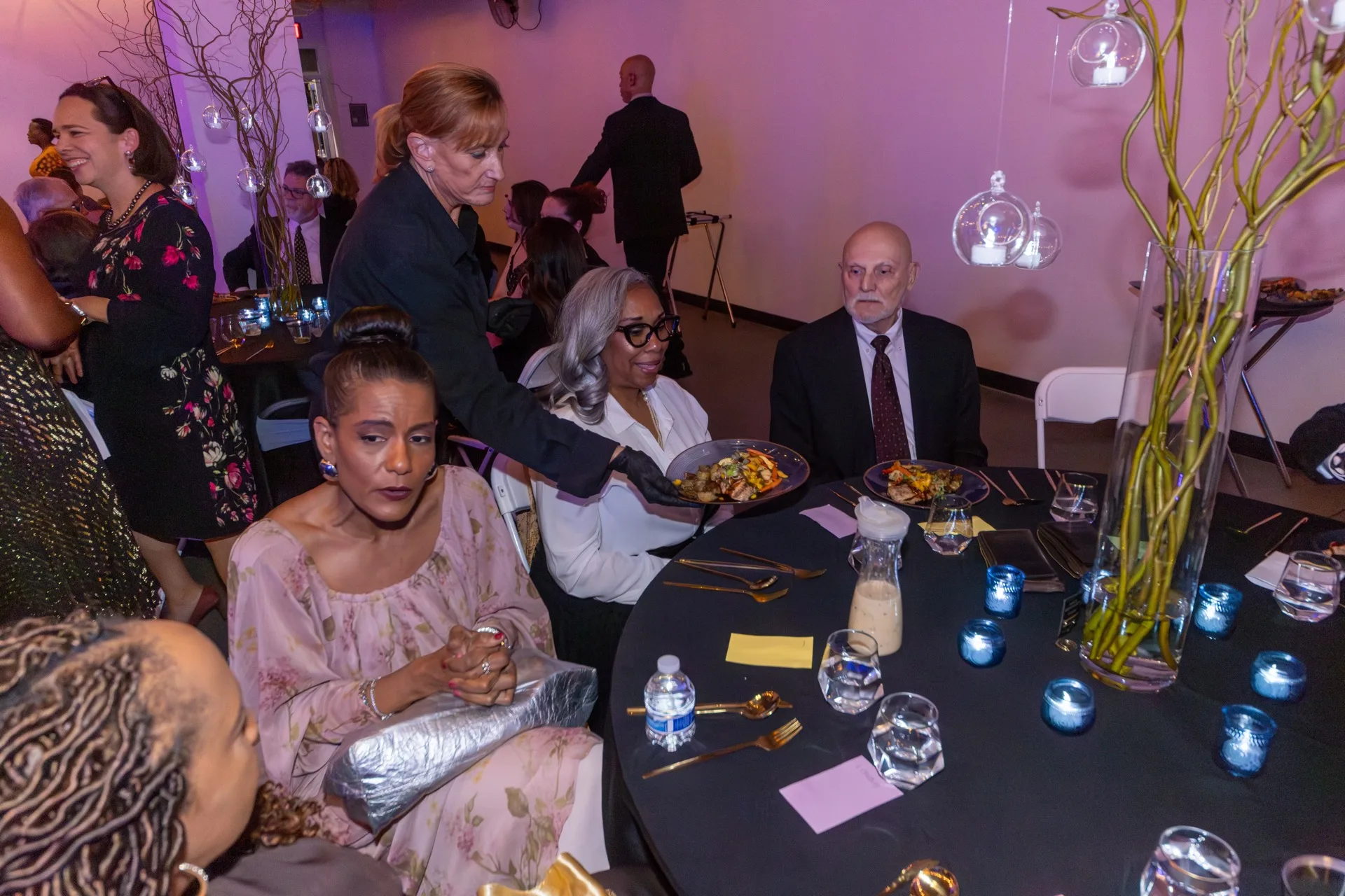 A server places a plated dinner in front of seated guests at a round table set with candles and glasses.