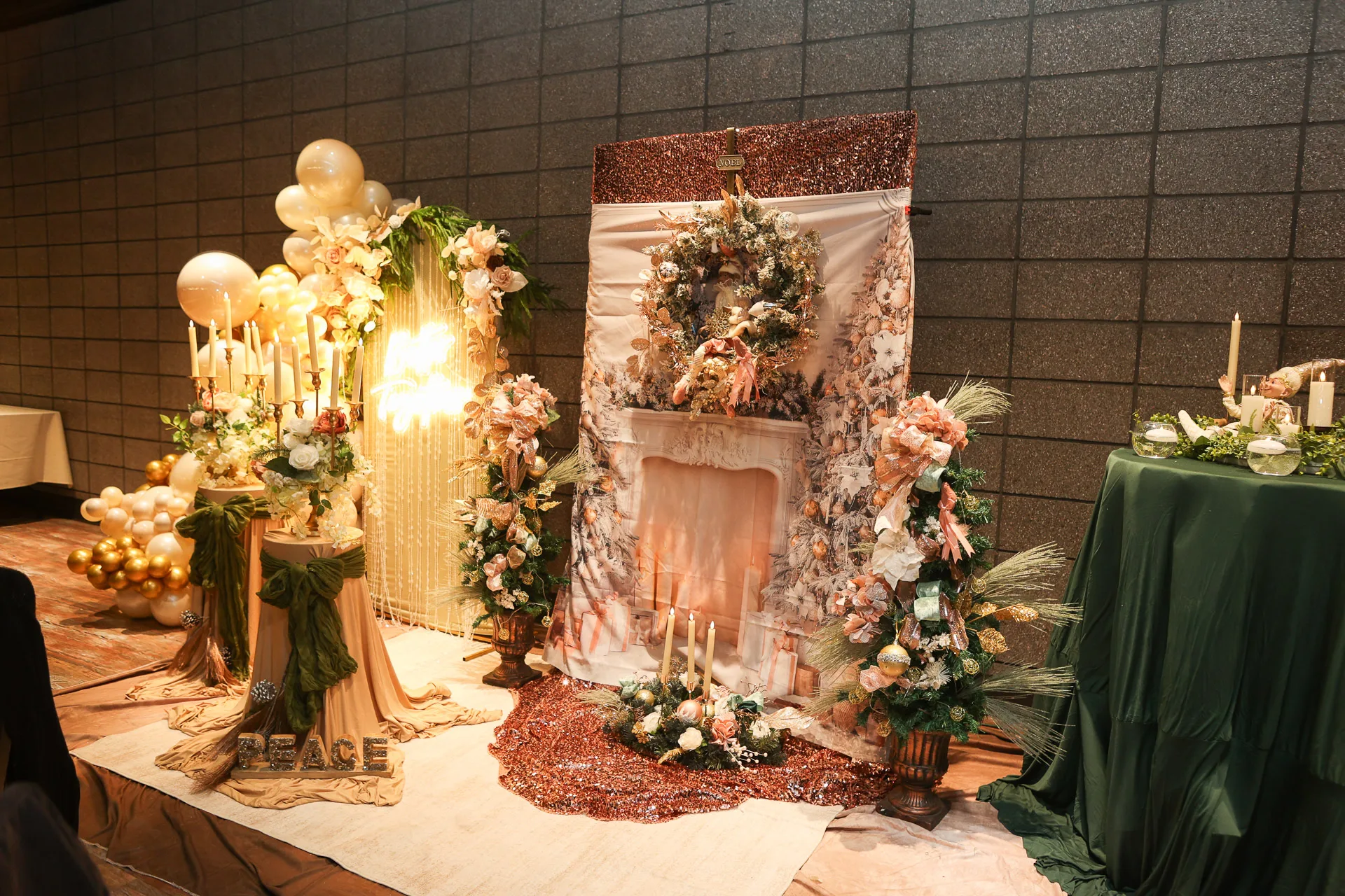 A decorated faux fireplace backdrop with a floral wreath, candles, balloon garlands, and a green-draped table beside it.