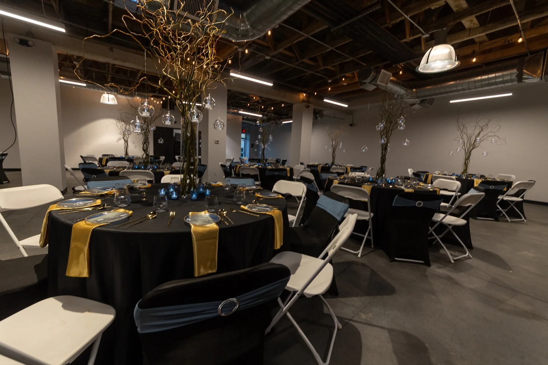 Formal event venue with round black-linen tables, gold accents, white chairs, tall branch centerpieces with hanging ornaments, and bright industrial overhead lighting.
