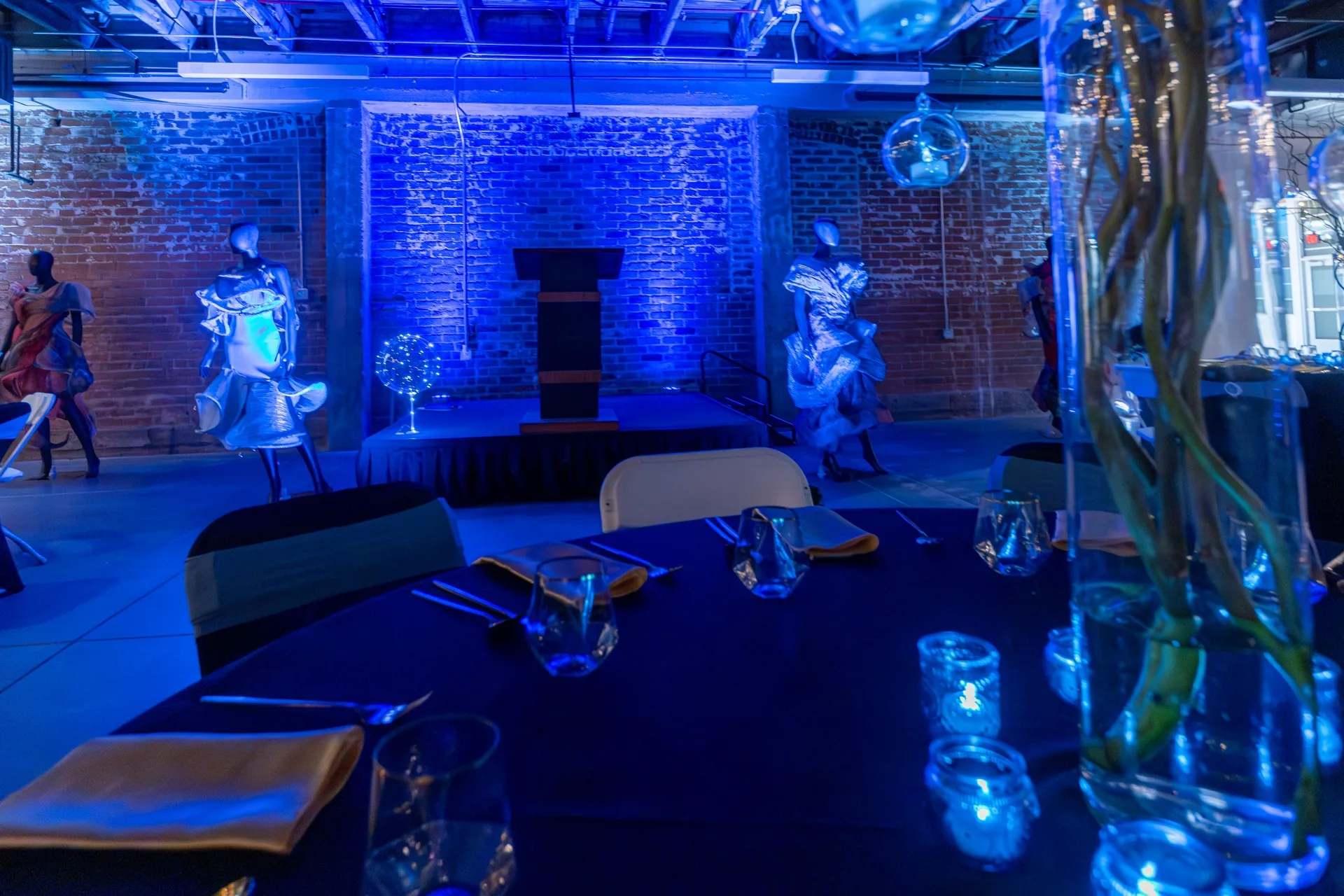 Event room with blue uplighting on brick walls, a lit stage podium, and a candlelit table setting in the foreground.