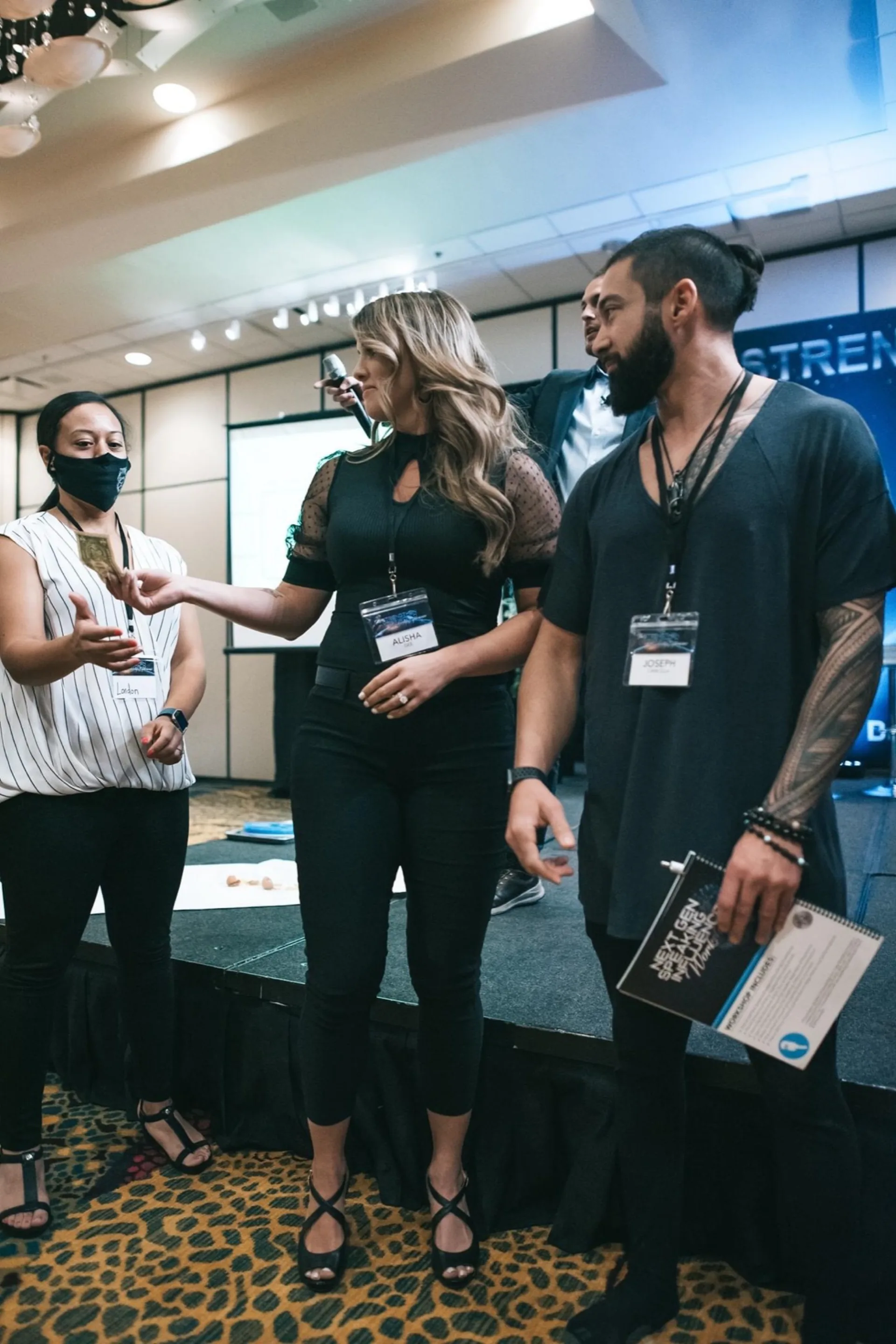 Four people at an indoor conference stage wear printed name badges on lanyards while one hands over a small item.