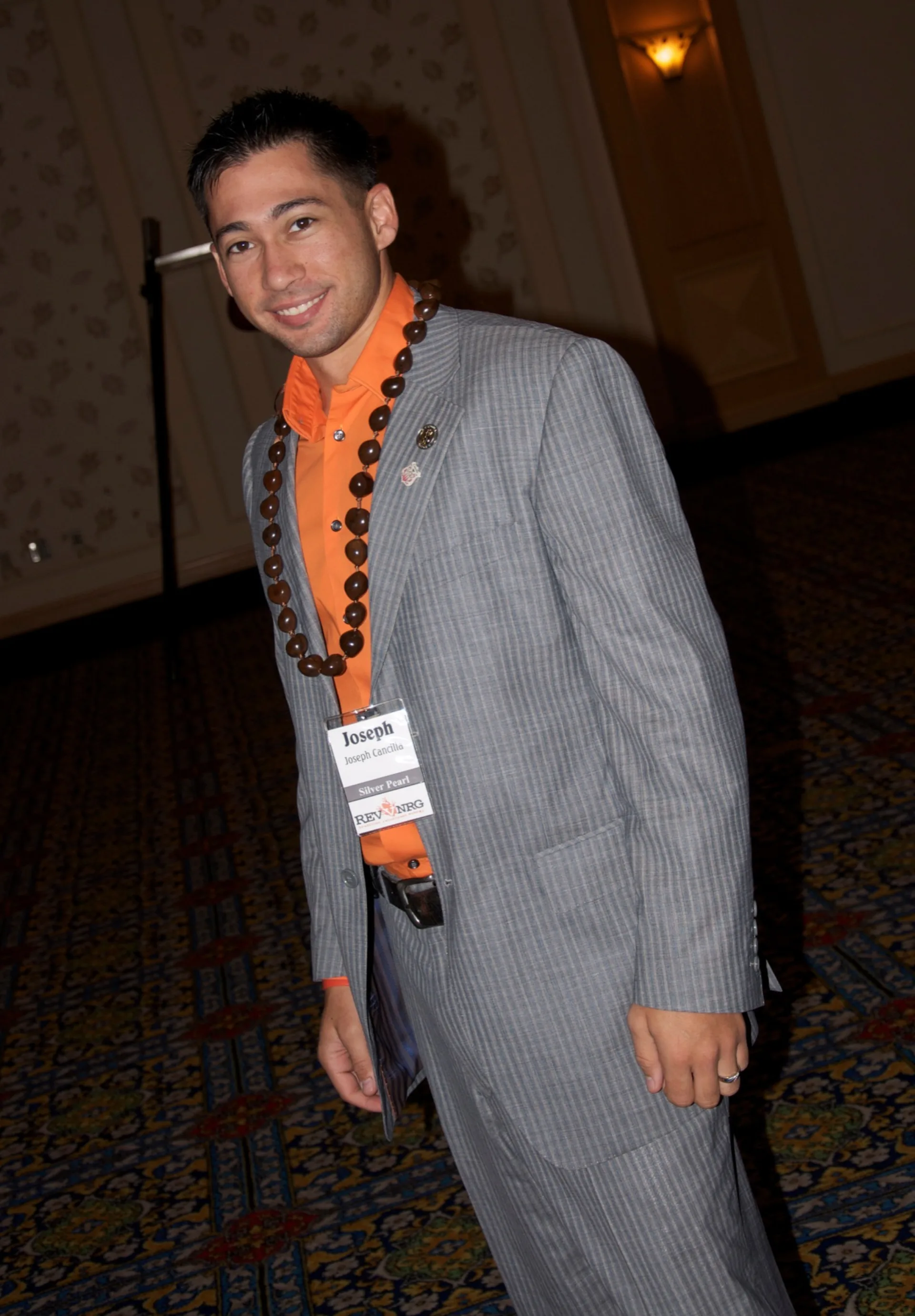 Man in a gray suit and orange shirt wears a printed name badge on a lanyard at an indoor event.