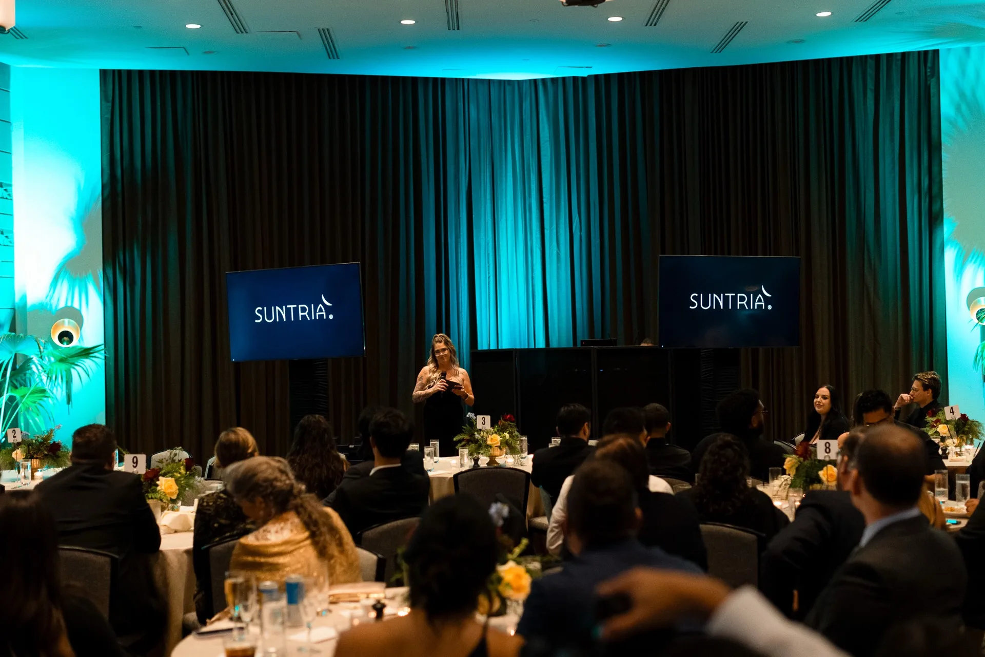 Corporate gala with teal uplighting, draped stage backdrop, and round tables set with floral centerpieces and white linens.