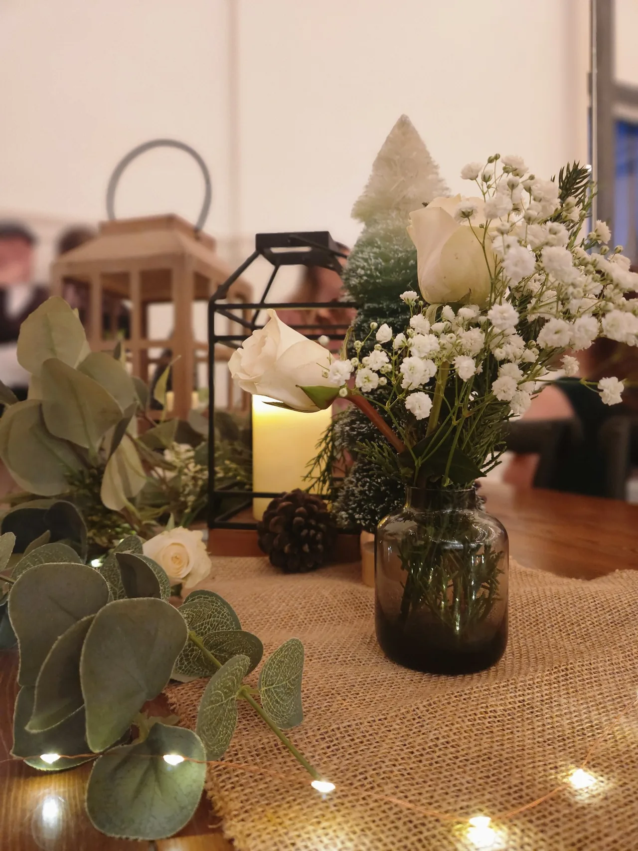 Close-up Hana event centerpiece with a lantern candle, white roses, eucalyptus greenery, and twinkling string lights on burlap