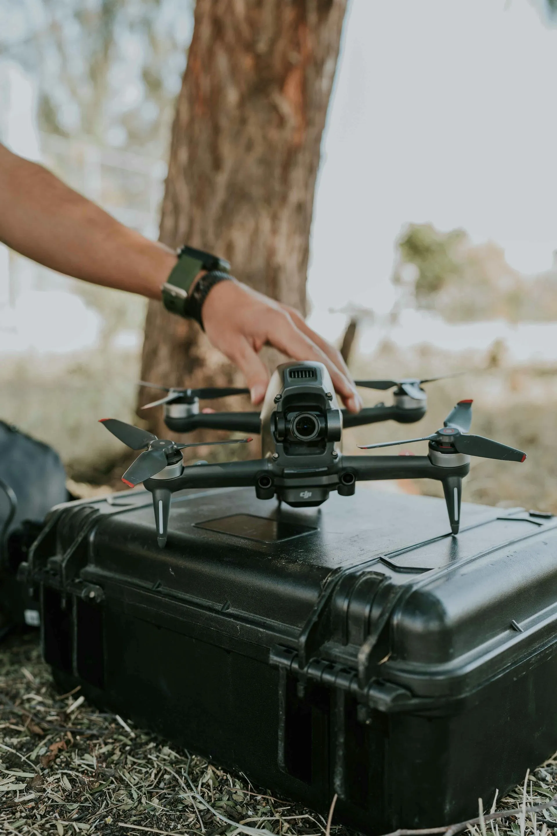 Drone on a black case, with a person's hand touching it. Outdoors near a tree.