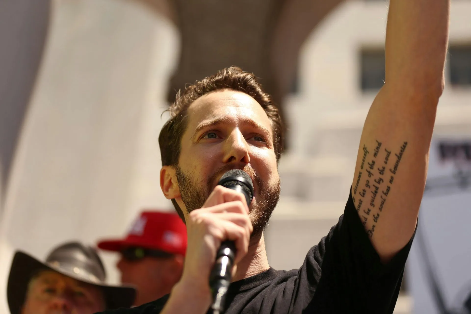 Man with a beard speaks into a microphone, arm raised, a tattoo visible on his upper arm.