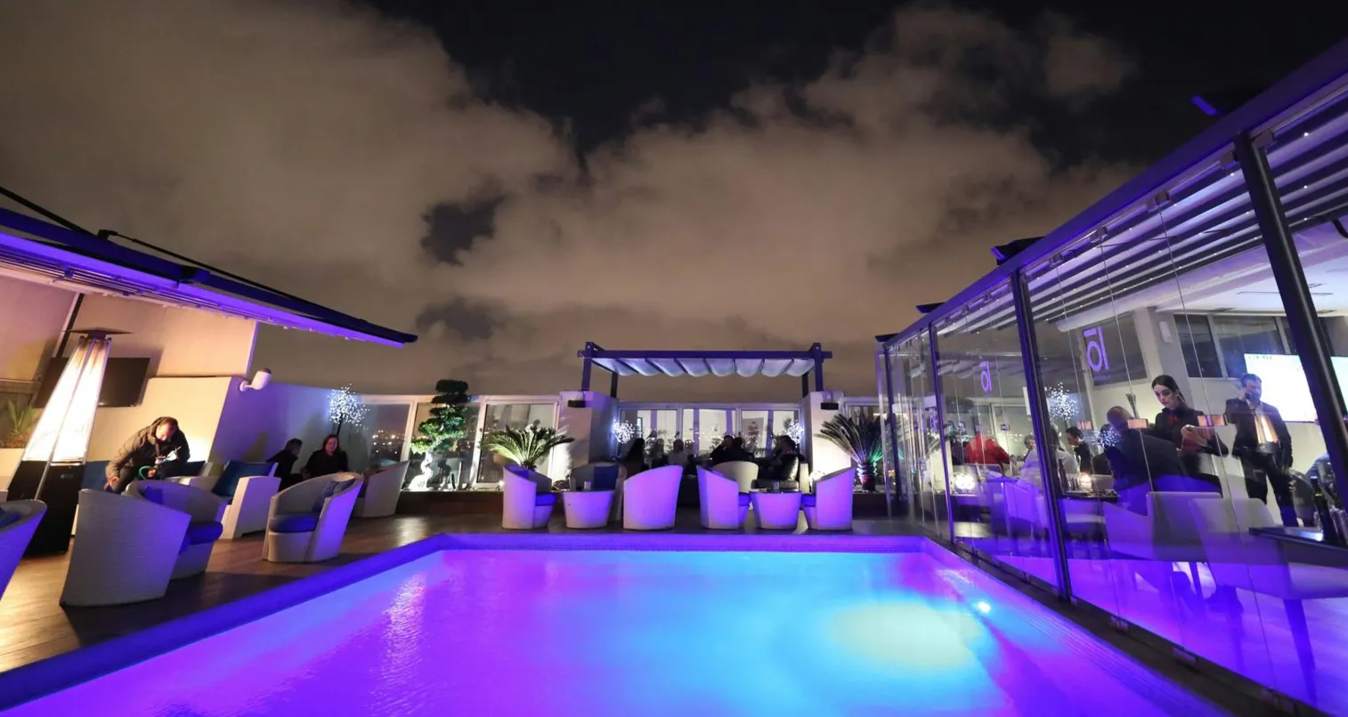 Rooftop pool and bar at night with people, white furniture, and purple lighting under a cloudy sky.