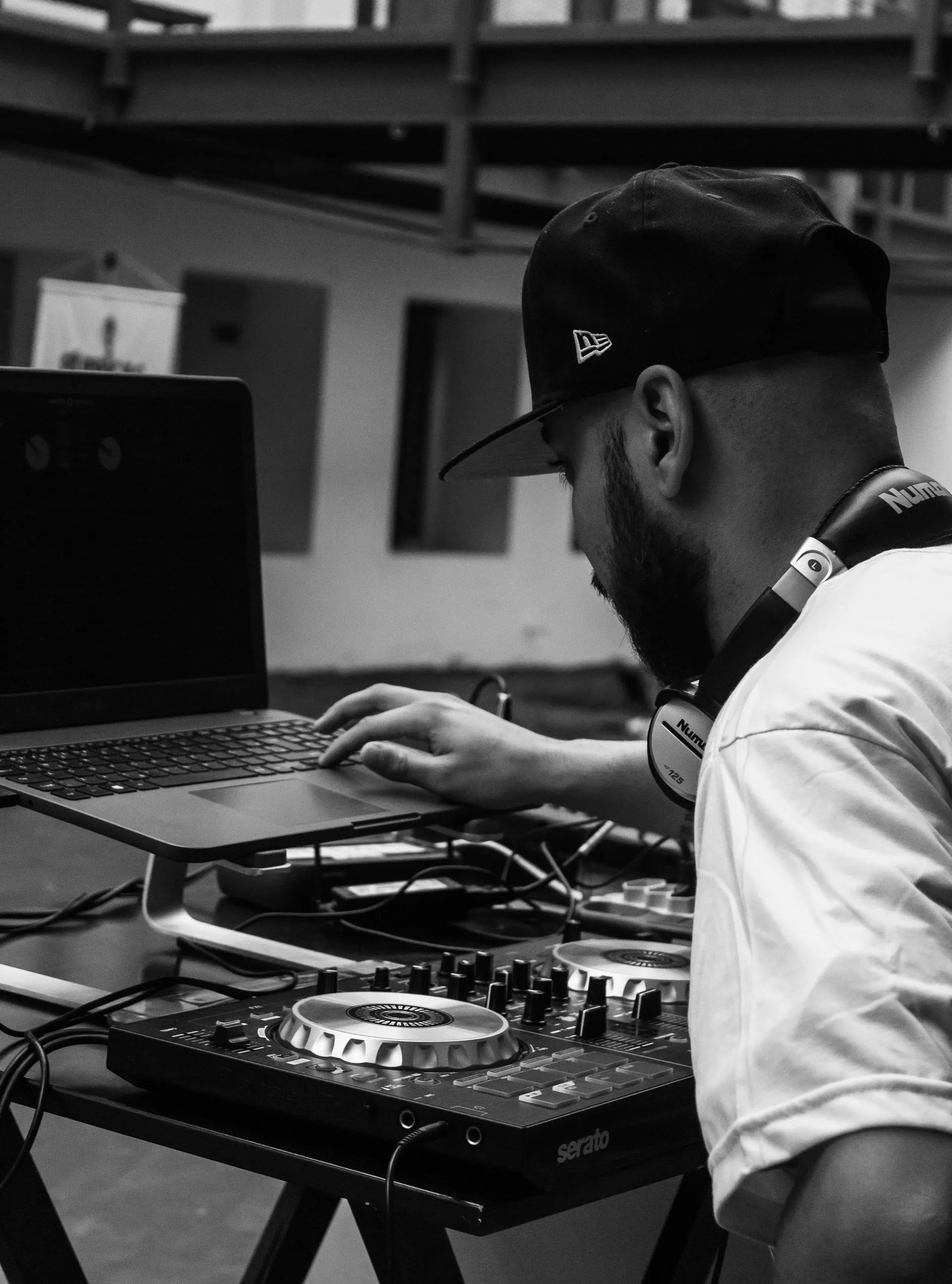 DJ wearing headphones and a hat, using a laptop and mixing console. Black and white.