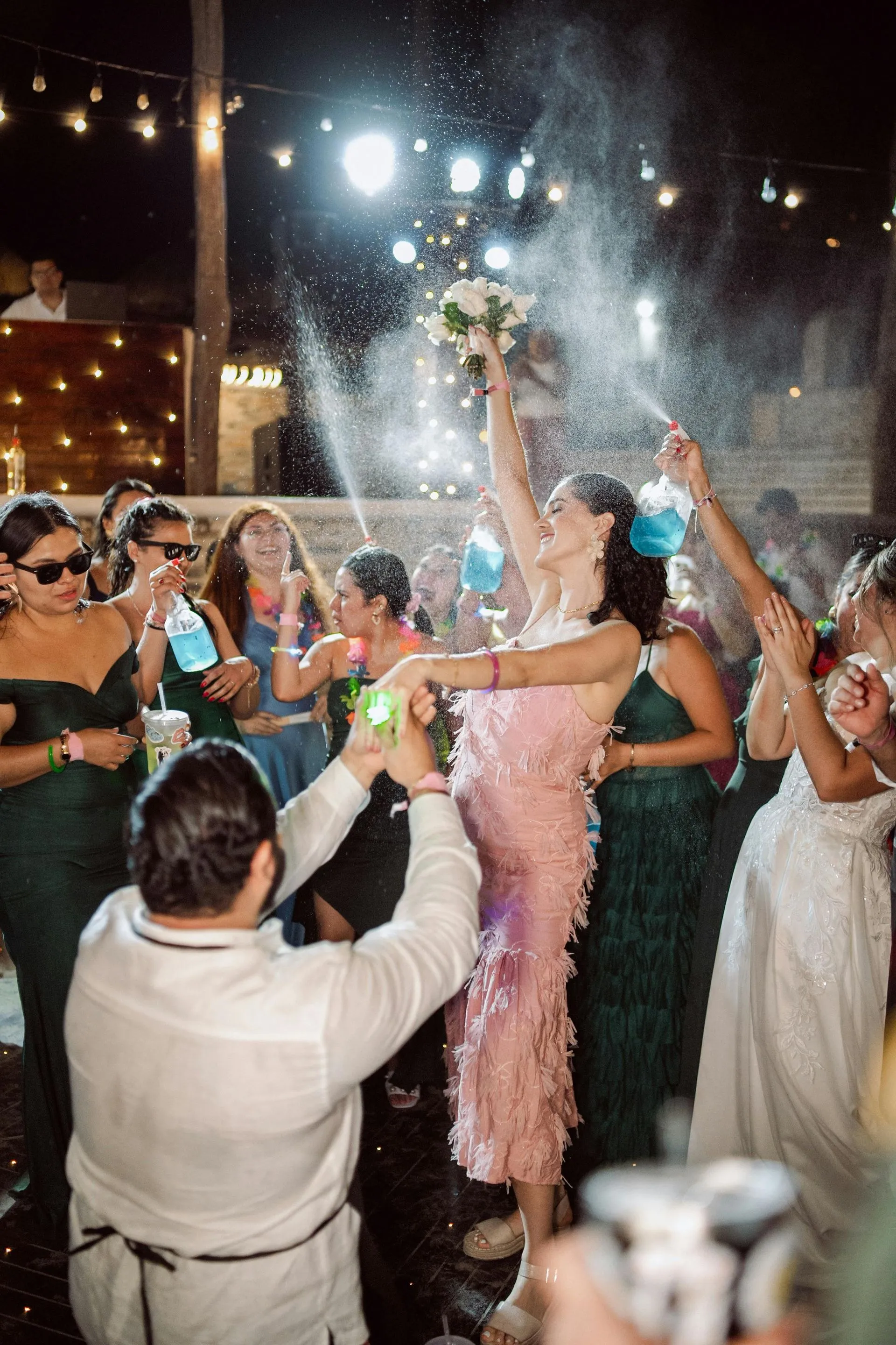 Wedding party celebrates with confetti, bride holding bouquet high. Night setting with lights.