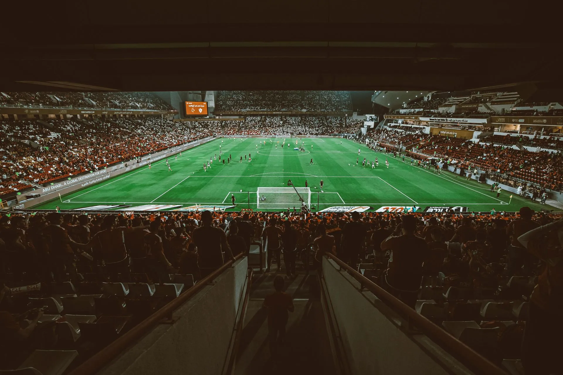 Soccer game in a packed stadium. Green field, white goalposts, red and white seating.
