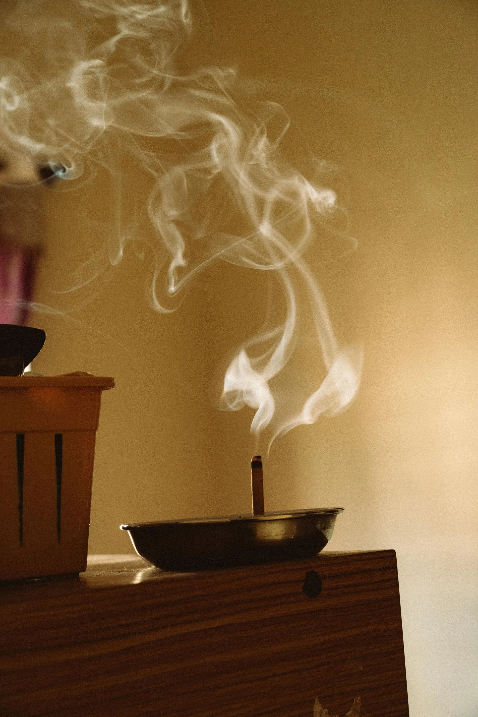 Incense stick burning in a small dish, with smoke rising against a tan wall, indoors.