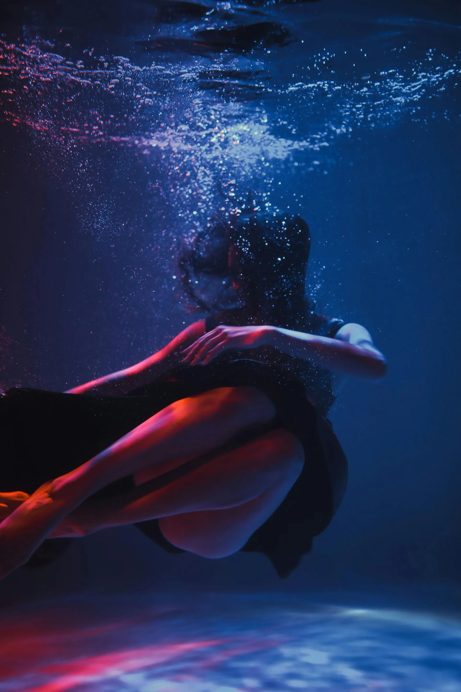 Woman underwater, black dress, illuminated by blue and red light, surrounded by bubbles.