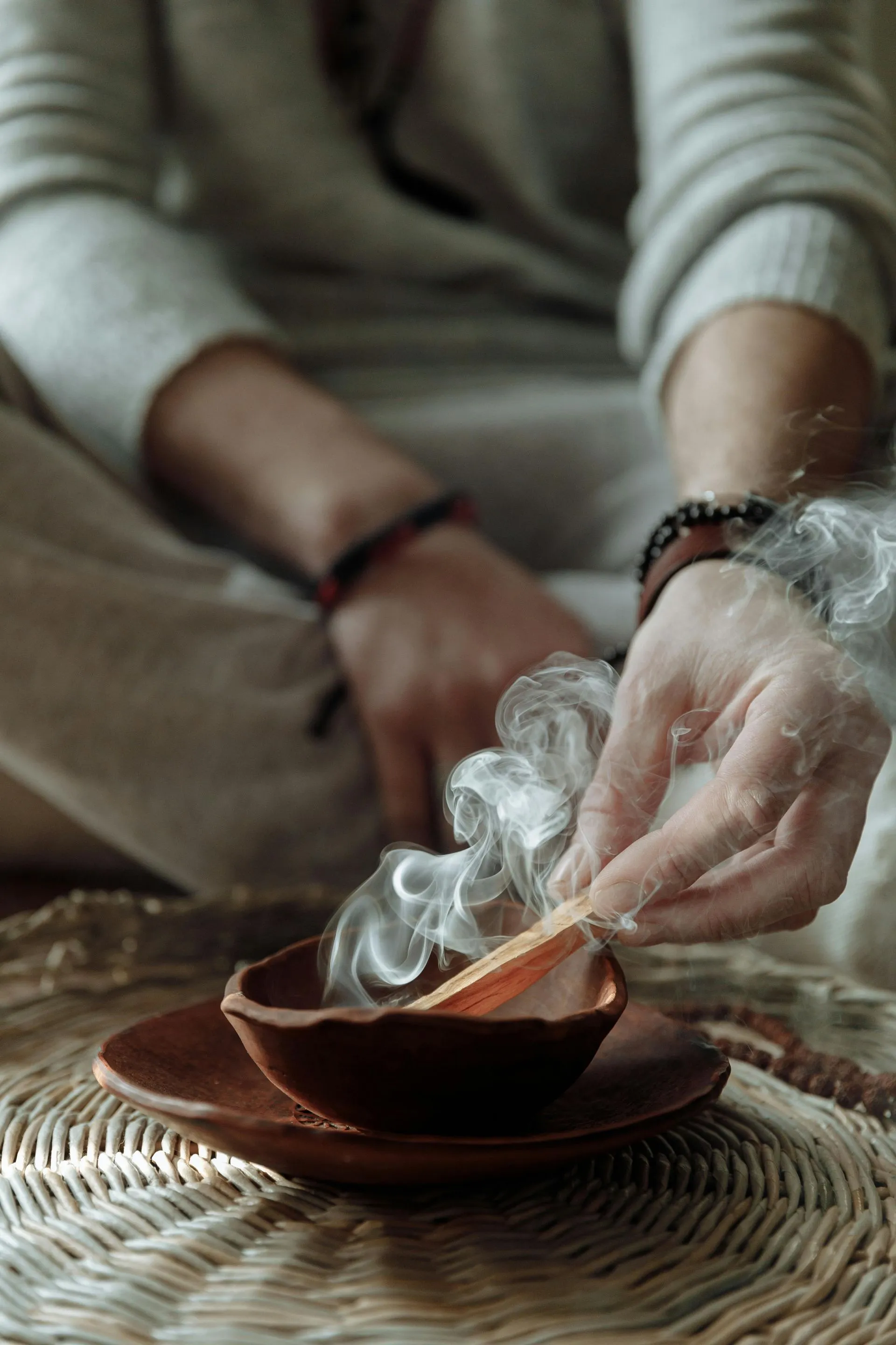 Person holding a smoking palo santo stick over a brown bowl. Smoke rises.