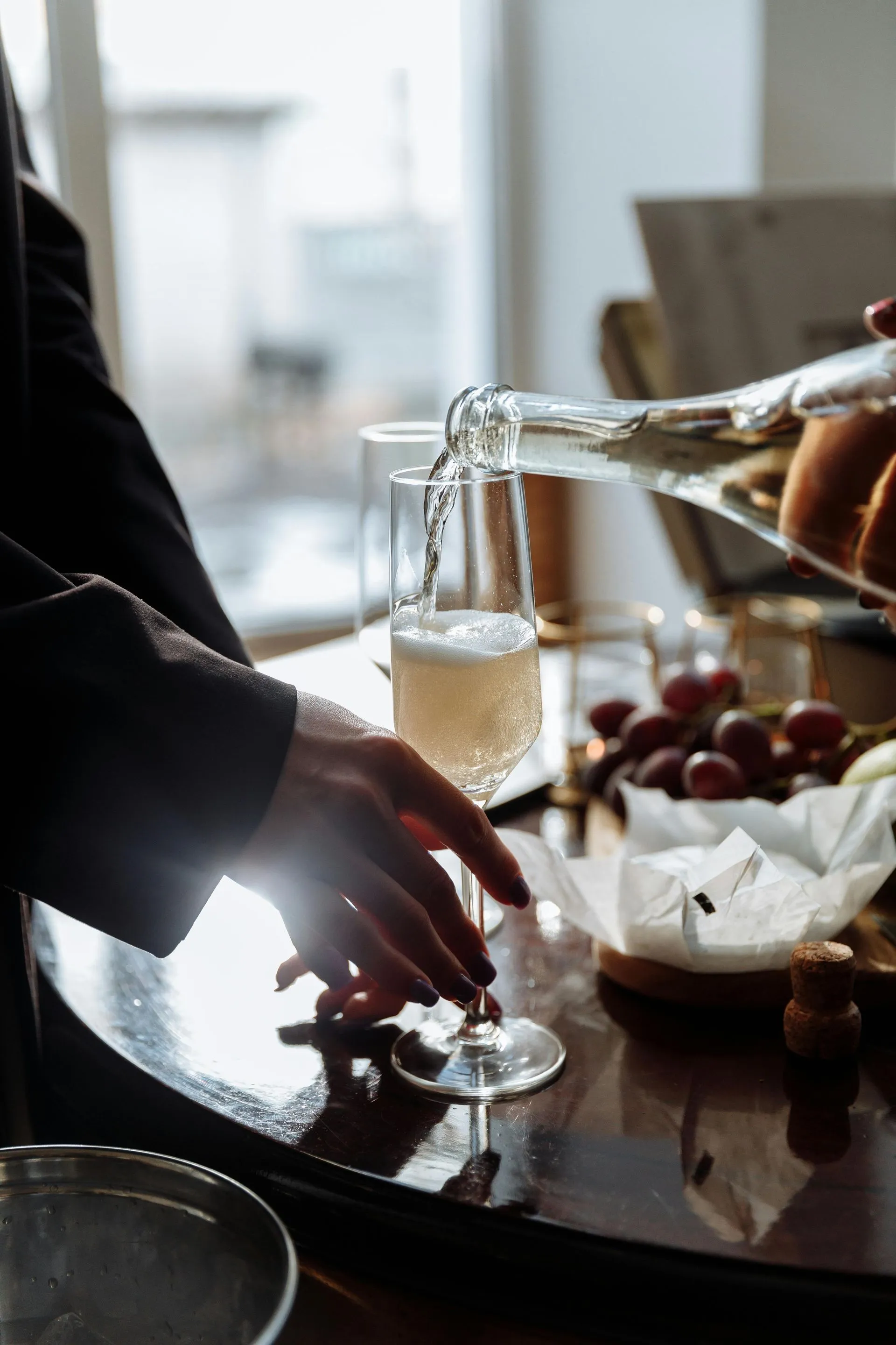 Champagne being poured into a tall glass at a table with grapes and snacks.