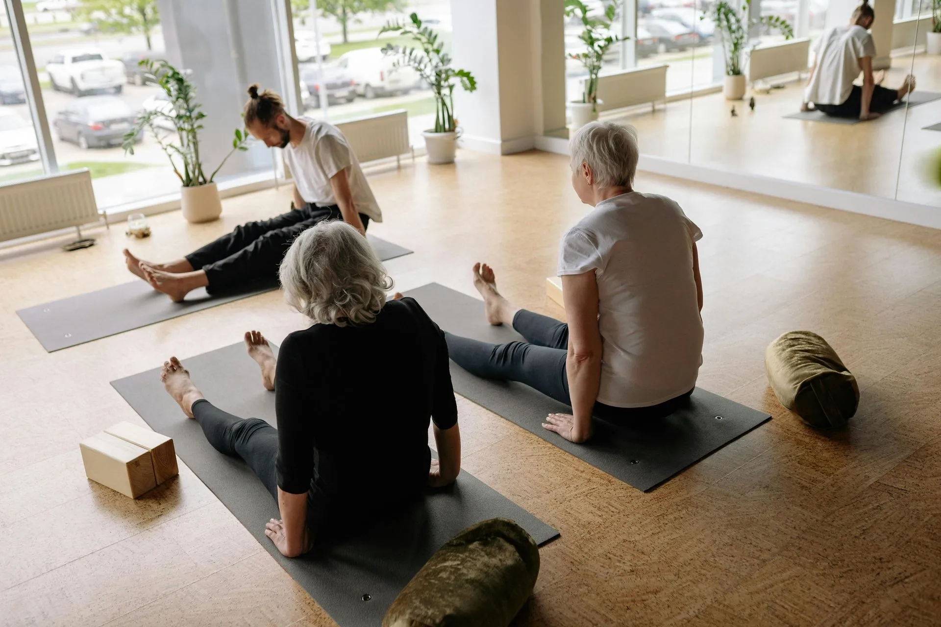 Yoga class: three people on mats, instructor in the background, a sunny room with windows.