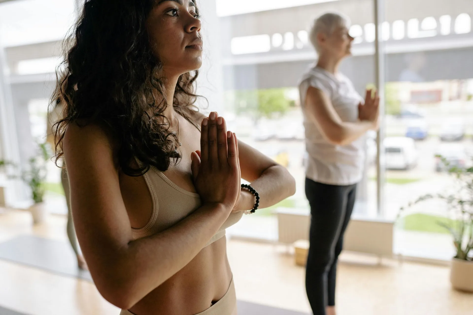Woman in yoga pose, hands in prayer position, facing upward in a bright room with another person in the background.